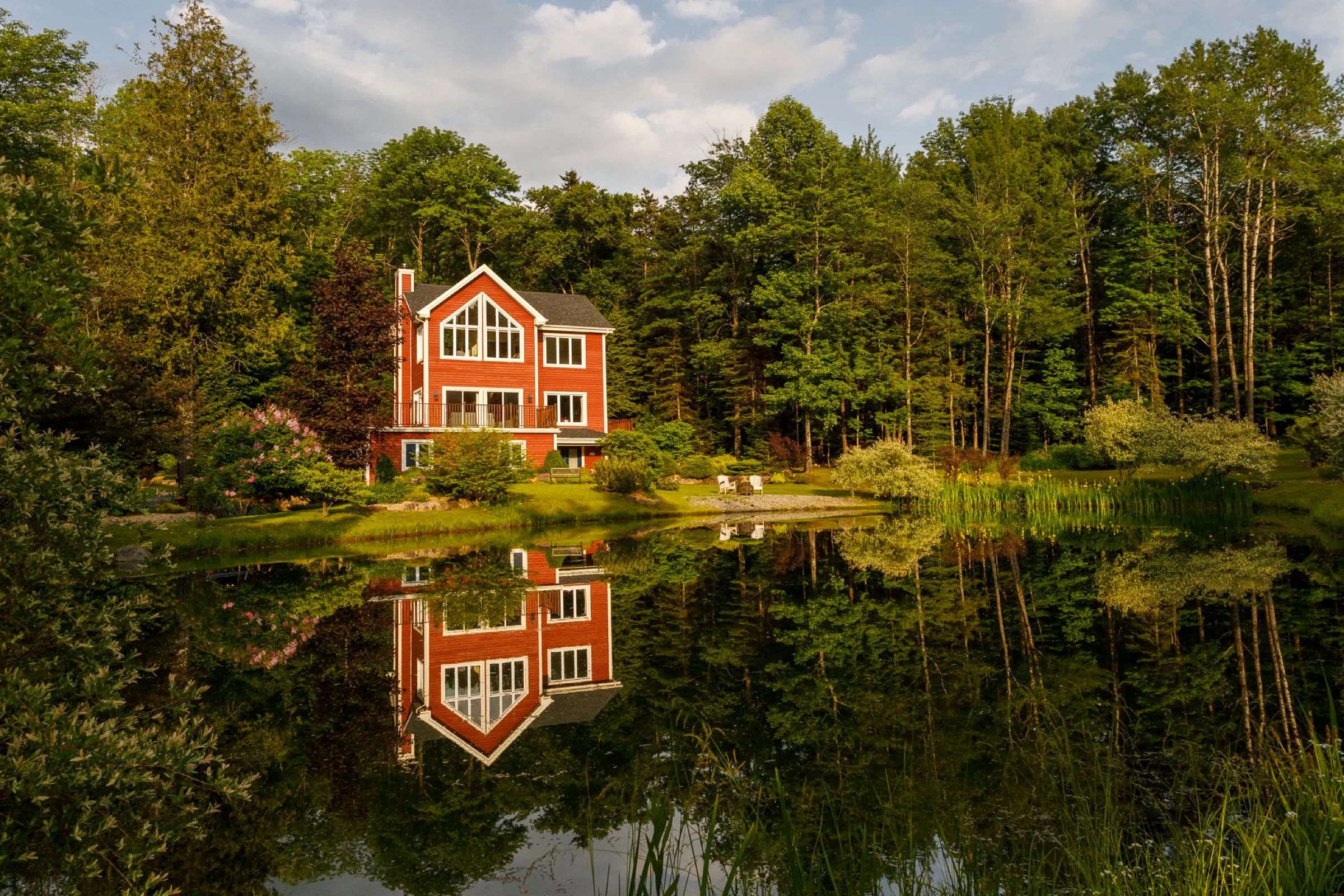 Maison de bois rouge en bordure de lac, entourée de conifères et réfléchie dans une eau immobile