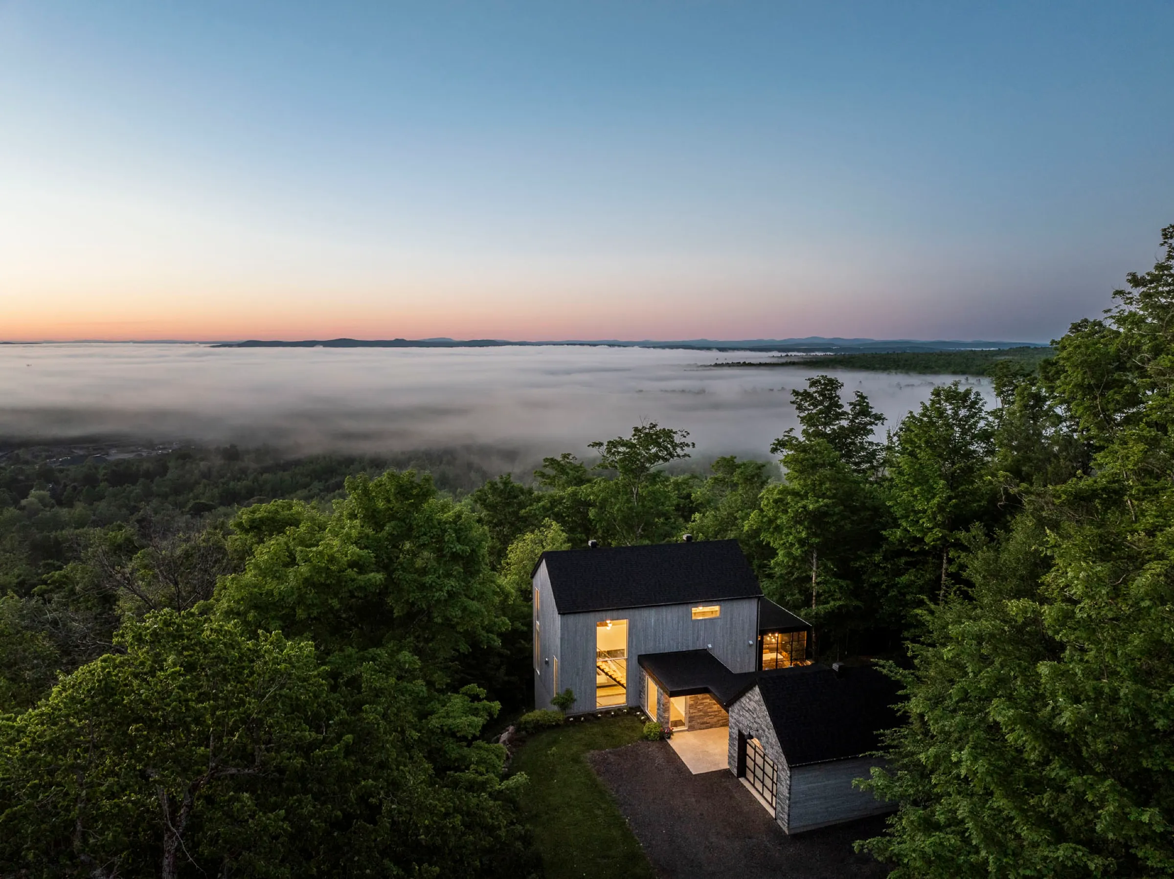 Maison sur un promontoire boisé vue du ciel à l'aube, la vallée en contrebas remplie de brume
