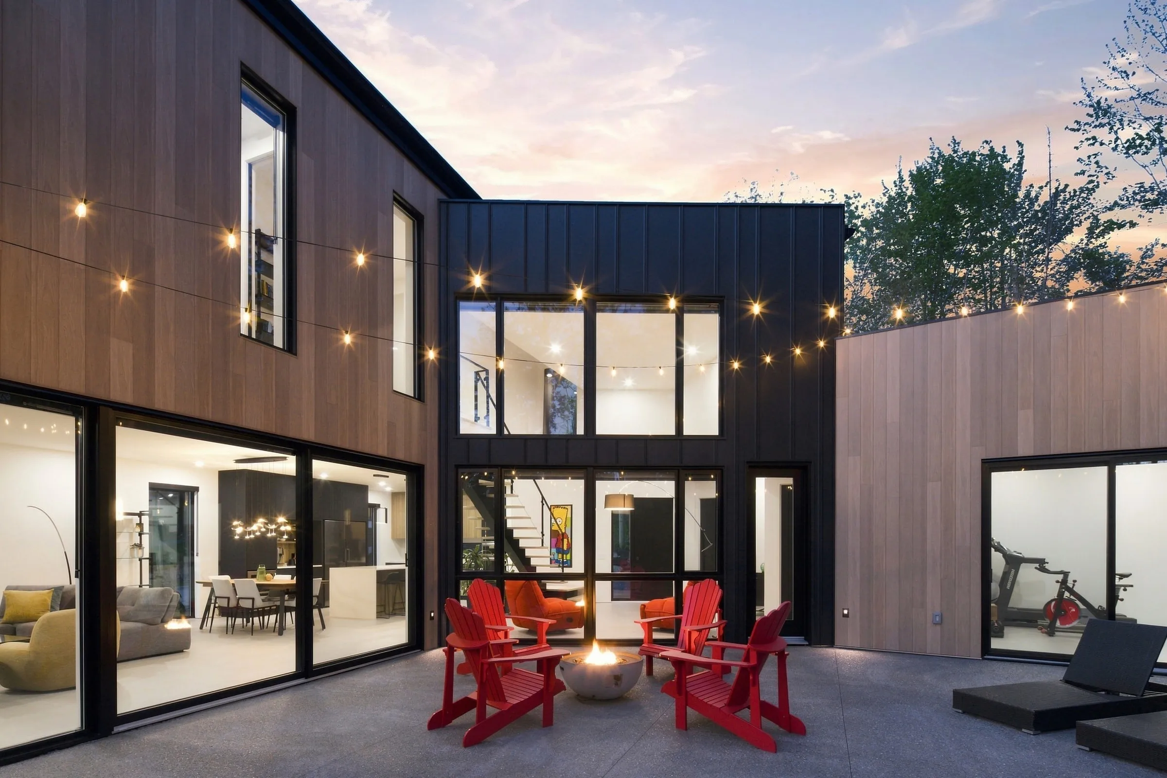 Maison contemporaine au revêtement noir, cour intérieure éclairée à la tombée du jour, fauteuils rouges devant la façade vitrée