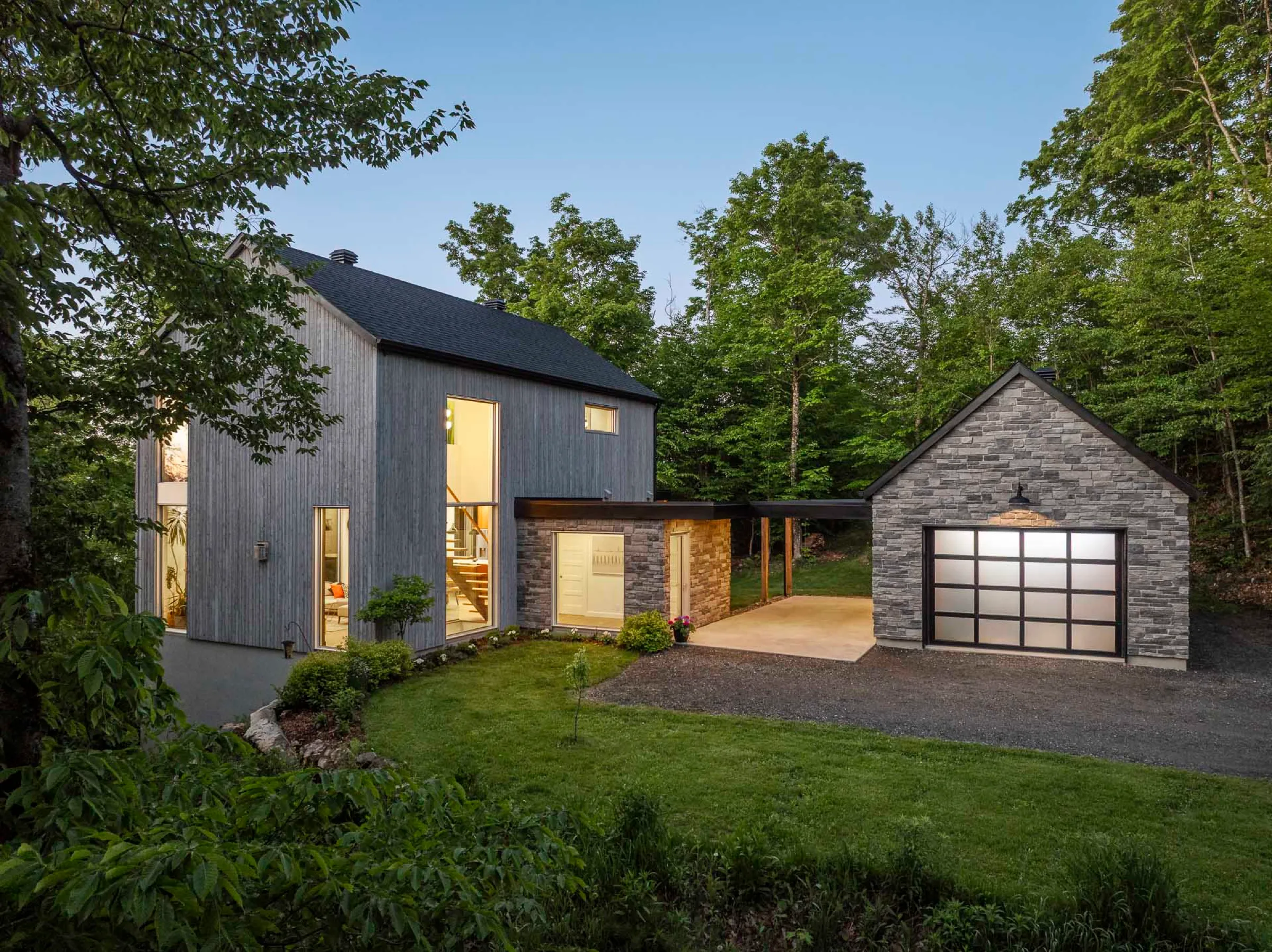 Façade contemporaine à revêtement gris et garage de pierre, pelouse dégagée, fenêtres éclairées en fin de journée