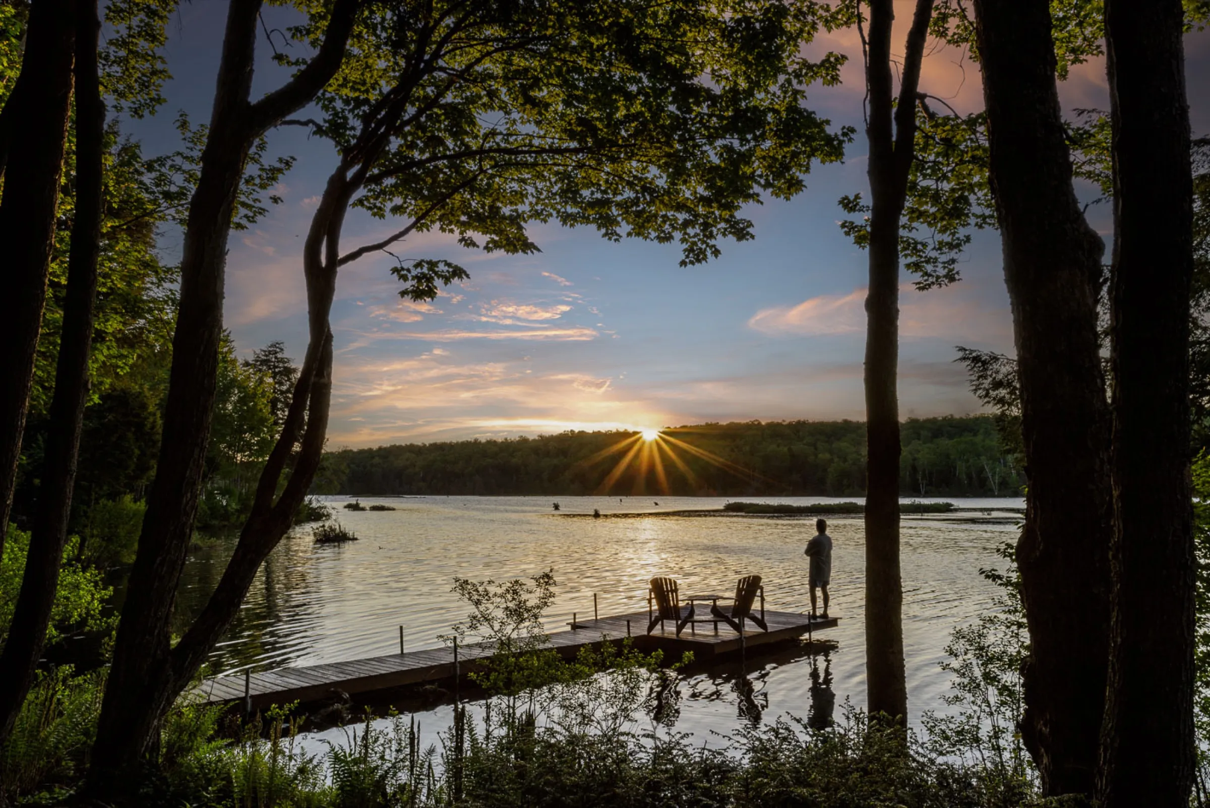 Quai de bois avançant sur un lac au coucher du soleil, vu depuis la rive entre les arbres