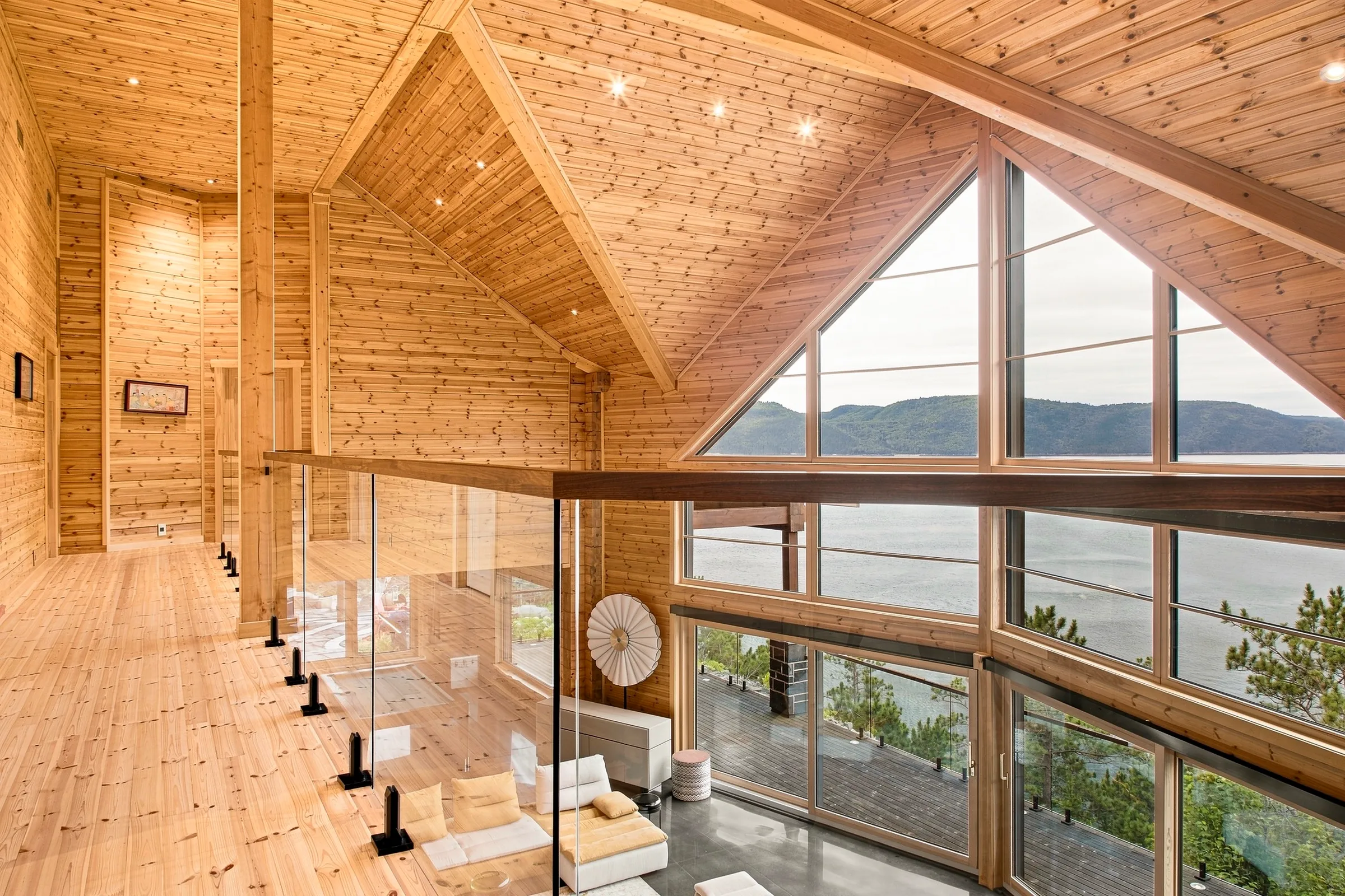 Mezzanine de bois sous charpente apparente, garde-corps de verre et vue plongeante sur le séjour