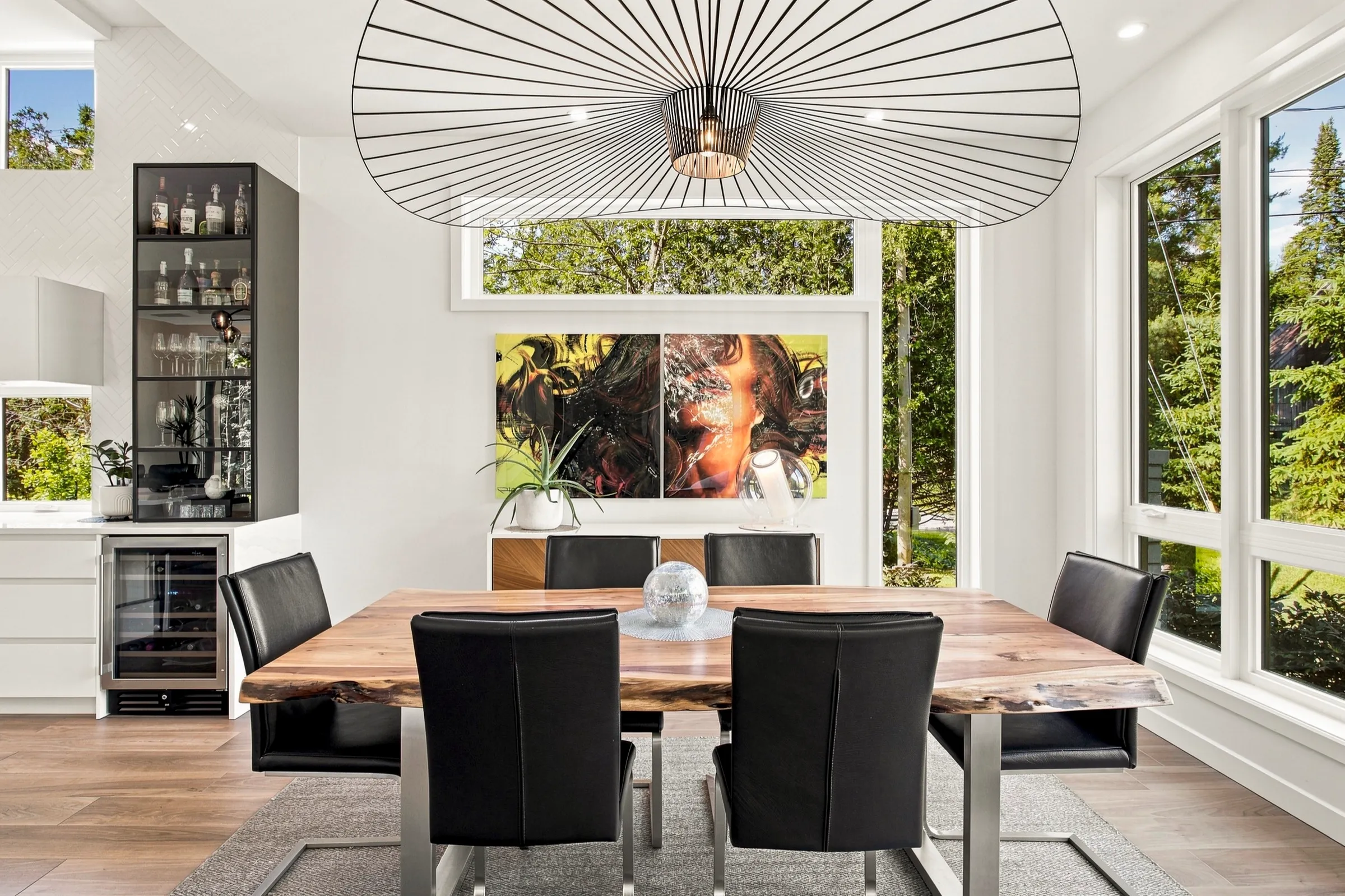 Salle à manger blanche sous un luminaire graphique, grande toile colorée et fenêtres sur le jardin