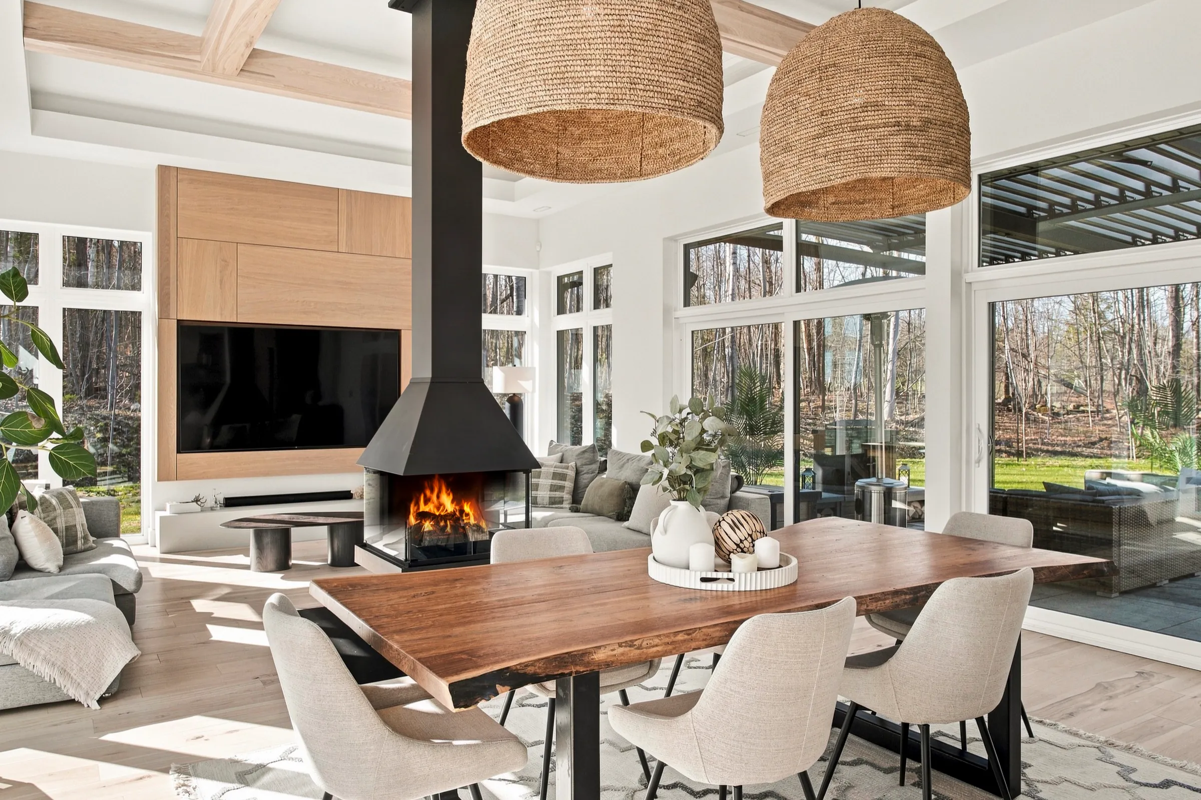 Table de bois sous suspensions tressées, foyer allumé et véranda vitrée en arrière-plan