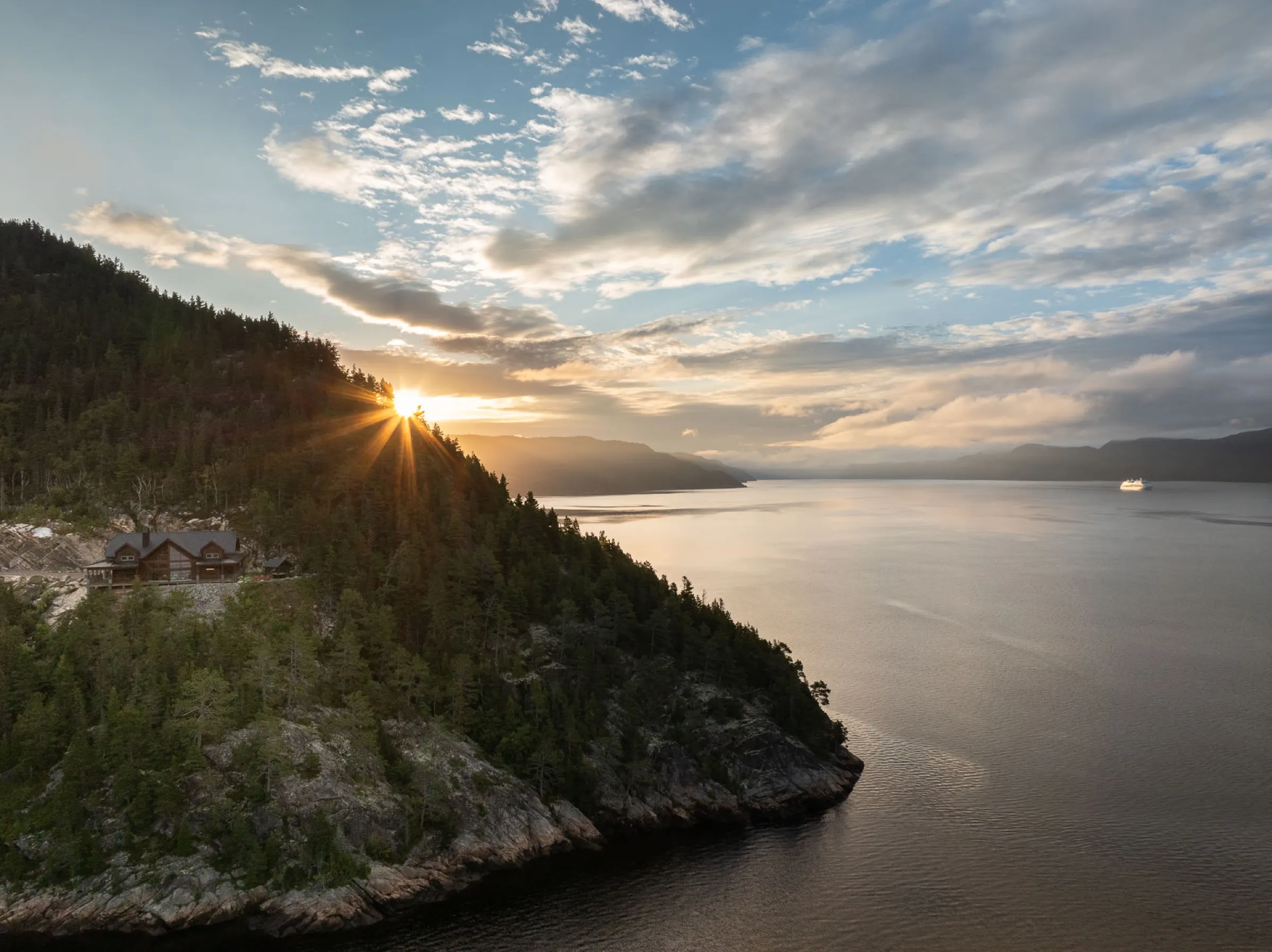 Chalet accroché au flanc d'une falaise boisée dominant le fjord au lever du soleil, un navire de croisière au loin sur l'eau