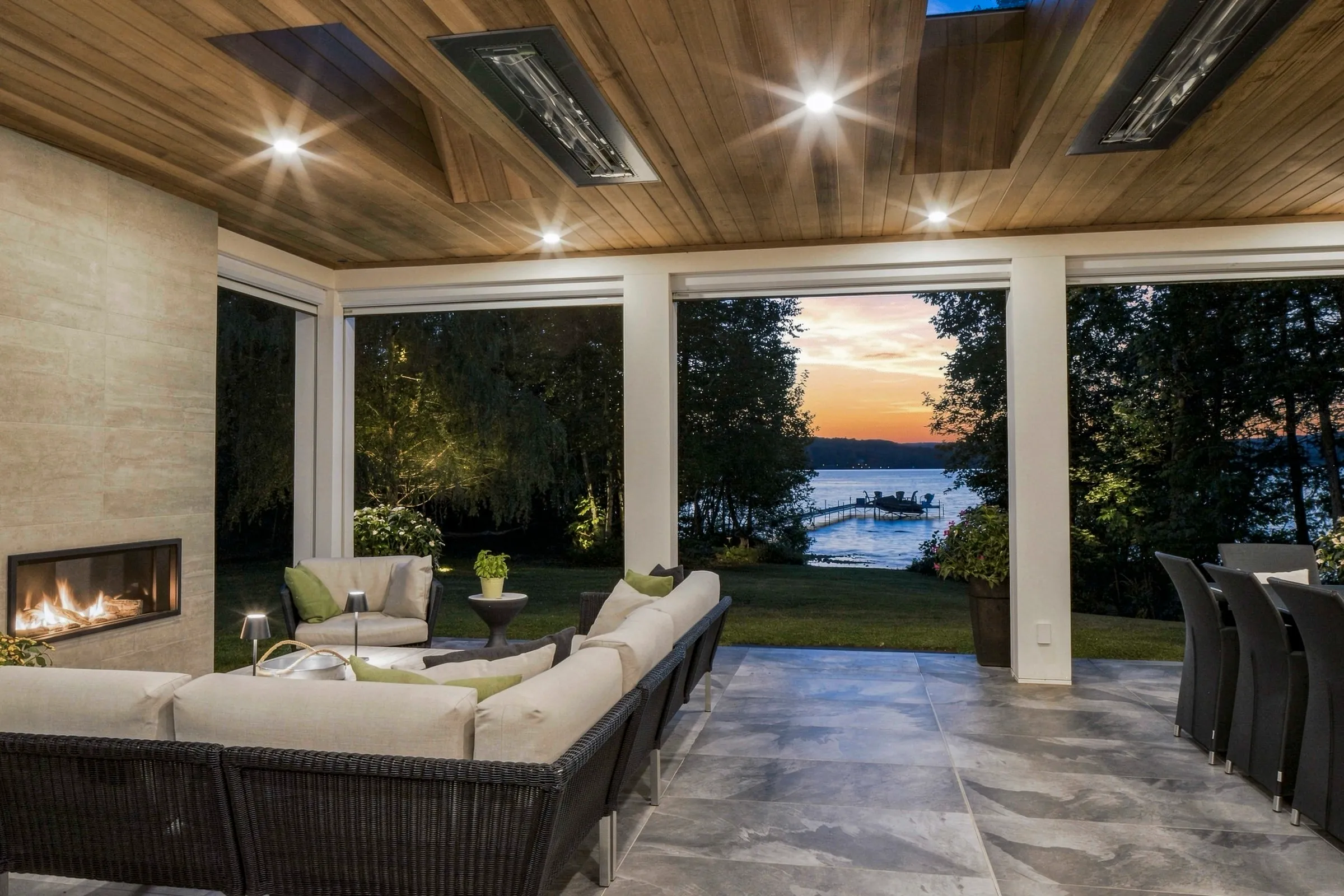 Terrasse couverte au plafond de bois, foyer linéaire allumé et mobilier de jardin face au lac à la tombée du jour