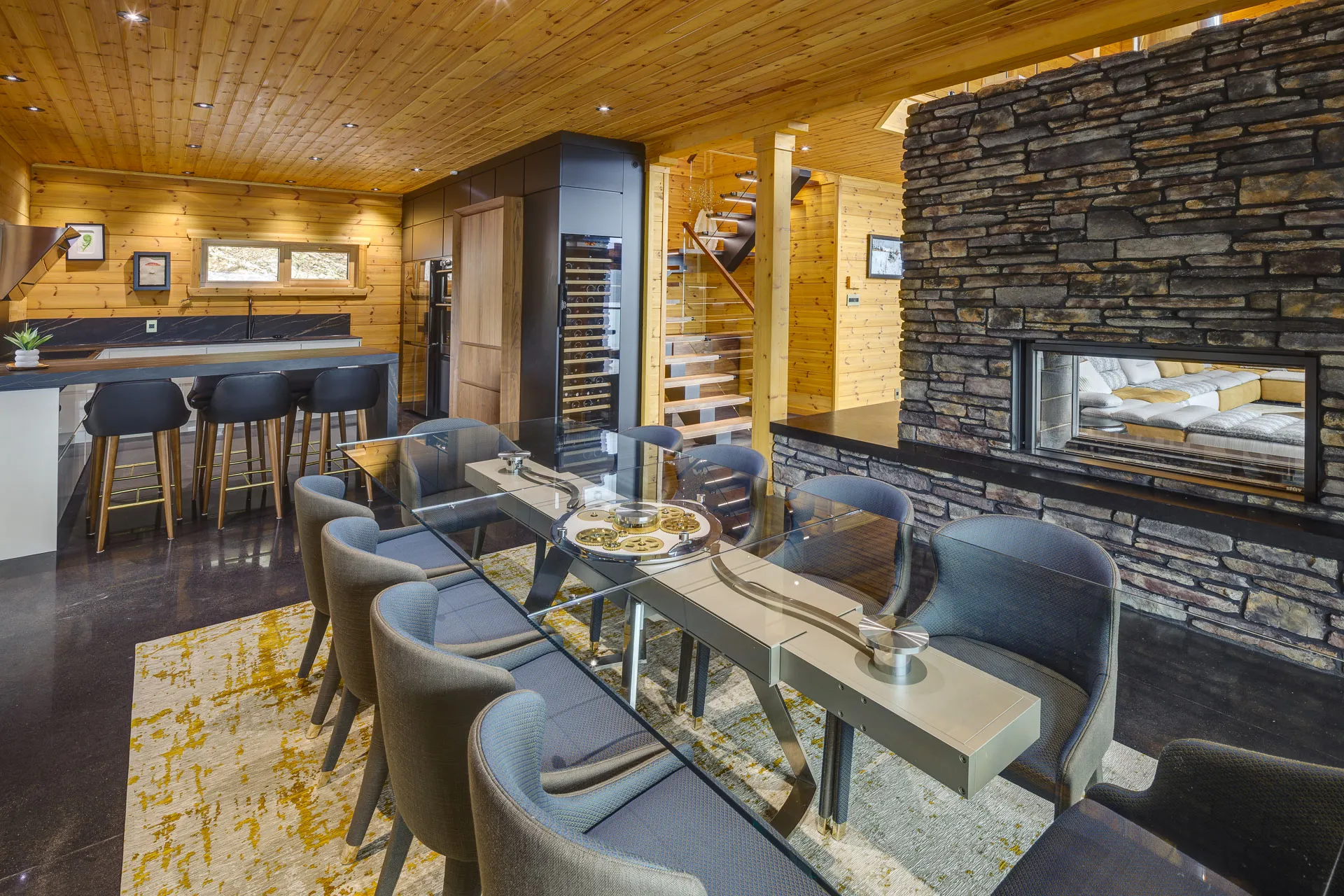 Salle à manger de chalet en bois, longue table de verre devant un foyer de pierre traversant qui ouvre sur le séjour, bar et cellier en arrière-plan